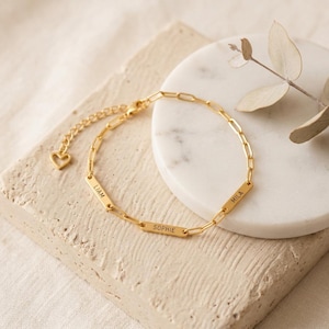 Puede incluir: Una pulsera dorada con un diseño de eslabones de cadena. La pulsera tiene tres placas rectangulares con los nombres "LIAM", "SOPHIE" y "MILA". Un pequeño dije de corazón está unido a la cadena ajustable.