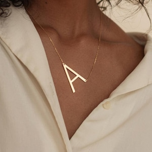 Può includere: Una collana color oro con un ciondolo a forma di lettera "A". Il ciondolo è una grande lettera maiuscola stilizzata sospesa a una catena sottile. La collana è indossata su una camicia di colore chiaro.