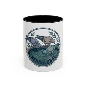 May include: White ceramic mug with a black interior rim. The mug features a circular design with three pigeons drinking water from a stone basin, with the text "HENAGLOBAL" below. Green leaves and a blue water design complete the illustration.