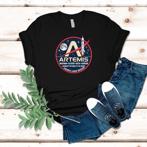 May include: Black t-shirt featuring the Artemis logo, a circular design with a rocket, moon, and stars. The text includes "ARTEMIS" and "HUMANITY RETURNS TO THE MOON CREWED LUNAR MISSION." The shirt is paired with blue jeans and sneakers.