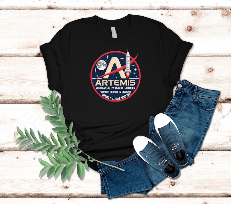 May include: Black t-shirt featuring the Artemis logo, a circular design with a rocket, moon, and stars. The text includes "ARTEMIS" and "HUMANITY RETURNS TO THE MOON CREWED LUNAR MISSION." The shirt is paired with blue jeans and sneakers.
