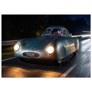 Puede incluir: Un coche plateado antiguo, con faros redondos y un diseño aerodinámico, se captura en movimiento por la noche. El coche tiene una matrícula que dice "T 2222". El conductor es visible dentro del vehículo. La imagen transmite velocidad y diseño automotriz clásico.
