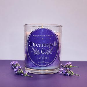 May include: A clear glass candle jar with a purple label featuring the text Persephone's Wick Co, Dreamspell, Scent Lavender, 2 oz, and Hand Poured Elegance. The label includes a crescent moon and star graphic. Small sprigs of lavender surround the base.
