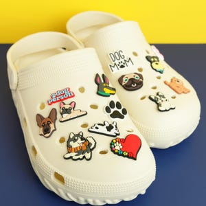 May include: A pair of off-white foam clogs decorated with various colorful rubber charms featuring dog breeds, paw prints, a red heart, and text that reads "dog person" and "DOG MOM" attached to the ventilation holes on the top surface.