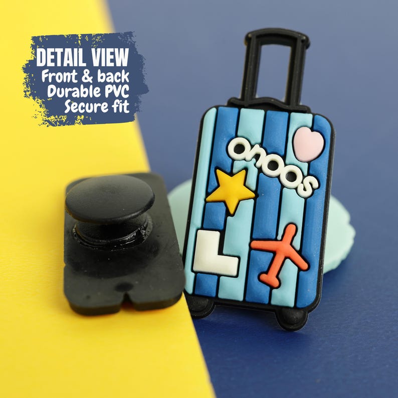 May include: A durable PVC shoe charm shaped like a blue and light blue striped suitcase. The charm features a yellow star, an orange airplane, a white letter L, a pink heart, and the text "onoos" in white. It includes a black circular attachment.