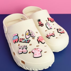 May include: A pair of off-white clogs decorated with colorful medical-themed charms. The charms include a stethoscope, lungs, a nurse's uniform, and the words "Nurse" and "Nurse Off Duty". The clogs are on a blue and pink surface.