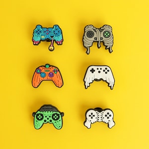 May include: A set of six decorative shoe charms shaped like video game controllers with various themes: a blue zombie controller with an eye, a grey mummy controller, an orange pumpkin controller, a white ghost controller, a green monster controller, and a white skull controller.