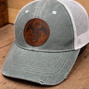 May include: A distressed gray baseball cap with a white mesh back. The cap features a round, brown leather patch with a dragon design. The bill has a distressed look with small tears.