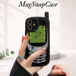 May include: Black phone case with a retro design, featuring a green screen with pixelated hearts. The case has a black antenna and silver buttons. The text "MagSnapCase" is at the top. The case is held by a hand with a checkered ring.