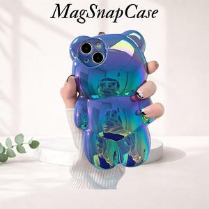Può includere: Una custodia per telefono blu iridescente a forma di orso è tenuta in mano. La custodia ha una finitura lucida e ritagli per la fotocamera del telefono. Il testo "MagSnapCase" è nella parte superiore dell'immagine.
