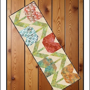 May include: A rectangular quilt runner titled Flowers for Mom by Villa Rosa Designs. The quilt features a pattern of stylized flowers in shades of red, orange, teal, and green on a cream background, with Expressions Batiks by Riley Blake Designs.