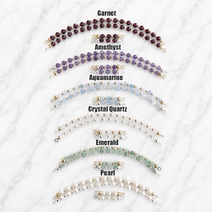 May include: A collection of gemstone bracelets in various colors, including garnet, amethyst, aquamarine, crystal quartz, emerald, and pearl. Each bracelet features small, round beads and gold-colored accents. The names of the gemstones are labeled above each bracelet.
