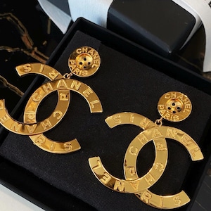 May include: A pair of gold-colored earrings in a black box. Each earring features a large, stylized "C" shape with the word "CHANEL" inscribed. A smaller circular element with "CHANEL PARIS" is attached to each earring.