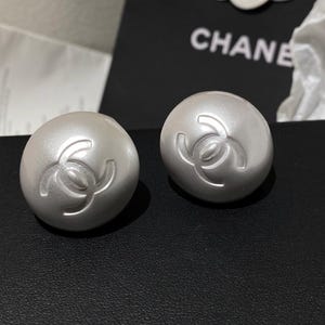 May include: A pair of pearl-like, round earrings. Each earring features a raised, interlocking double-C logo. The earrings are a light, pearlescent color and appear to be made of a smooth material. The word "CHANE" is visible in the background.
