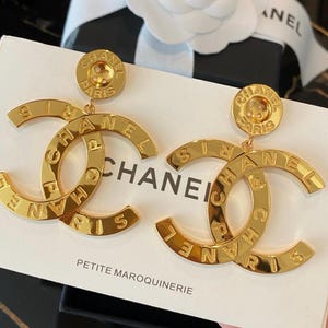 May include: A pair of gold-toned earrings. Each earring features a large, stylized 'C' shape with the word 'CHANEL' inscribed. The earrings have a button-like top with the words 'CHANEL PARIS' engraved. The earrings are on a white surface with the words 'CHANEL' and 'PETITE MAROQUINERIE' visible.