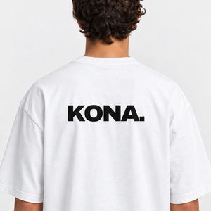 Può includere: Una t-shirt bianca a maniche corte con la parola KONA. stampata in grassetto nero e lettere maiuscole sulla parte superiore della schiena. Il capo presenta un girocollo e una vestibilità ampia, con il testo centrato orizzontalmente sul retro.