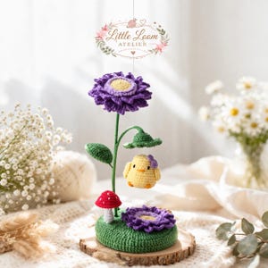 Op de afbeelding: Een handgemaakte gehaakte bloemstuk met een grote paarse bloem met een gele kern, een kleine gele kuiken met een paarse strik en een rode paddenstoel. De opstelling staat op een houten voet met een groene gehaakte basis.