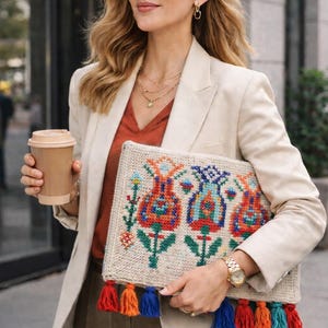 May include: A beige clutch with a colorful floral design and tassels. The clutch is held by a person wearing a cream blazer, orange top, and sunglasses. The clutch has a woven texture and a vibrant, embroidered pattern.
