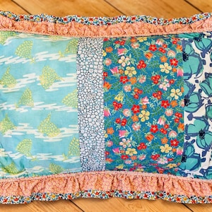 May include: Rectangular cushion with a patchwork design. It features sections of light blue fabric with sailboats, white fabric with small dots, teal fabric with red and yellow flowers, and teal floral fabric. A ruffled floral border frames the cushion.