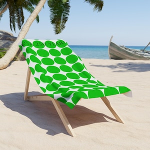 Puede incluir: Una silla de playa blanca con una toalla de playa con estampado ovalado verde y blanco. La toalla tiene el texto "USA 250 YEARS" impreso. La silla está en una playa de arena con un barco al fondo.