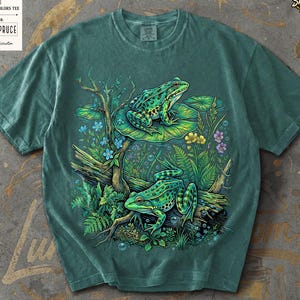 May include: Blue Spruce Comfort Colors tee featuring a vibrant illustration of two green frogs amidst lush aquatic plants and flowers. The design includes the text "Comfort Colors Tee" and "Blue Spruce" on a tag.