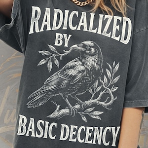 Puede incluir: Camiseta gris oscuro con las palabras "RADICALIZED BY BASIC DECENCY" en blanco. Una ilustración detallada en blanco y negro de un cuervo está en el centro. La camiseta está etiquetada como "COMFORT COLORS TEE" y el color es "PEPPER".