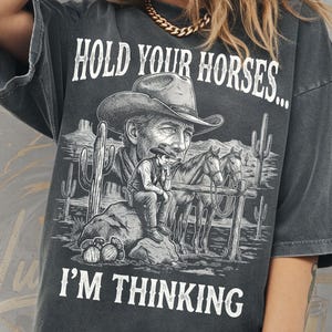 May include: Dark gray t-shirt with a cowboy graphic. The shirt features the text "HOLD YOUR HORSES... I'M THINKING" above and below a detailed illustration of a cowboy, horses, and desert scenery. The shirt is a comfort colors tee.