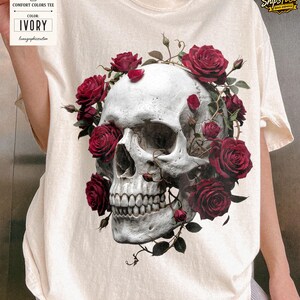 May include: Ivory-colored Comfort Colors tee featuring a detailed skull design adorned with vibrant red roses and green vines. The graphic is centered on the shirt, creating a striking contrast against the light background. The tag reads "COLOR: IVORY".