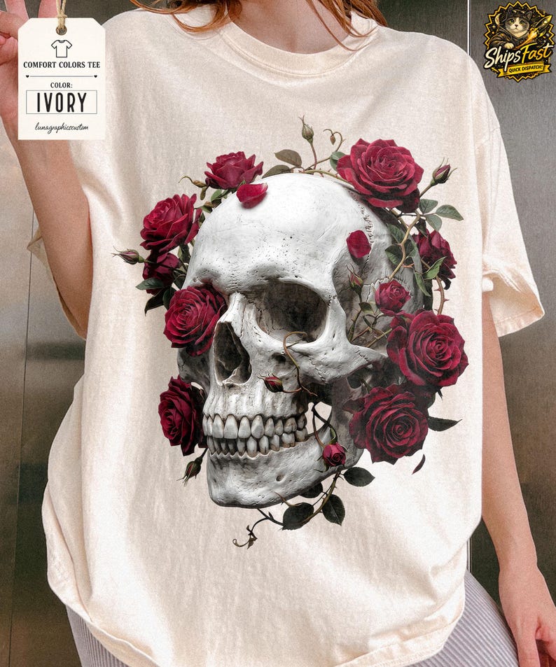 May include: Ivory-colored Comfort Colors tee featuring a detailed skull design adorned with vibrant red roses and green vines. The graphic is centered on the shirt, creating a striking contrast against the light background. The tag reads "COLOR: IVORY".