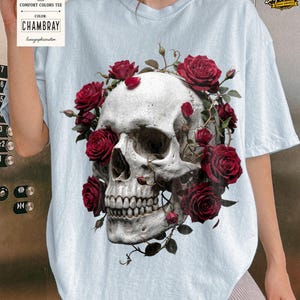May include: Light blue Comfort Colors tee featuring a detailed skull design encircled by vibrant red roses. The graphic tee includes the text "CHAMBRAY" on a tag. The design is a blend of gothic and floral elements.