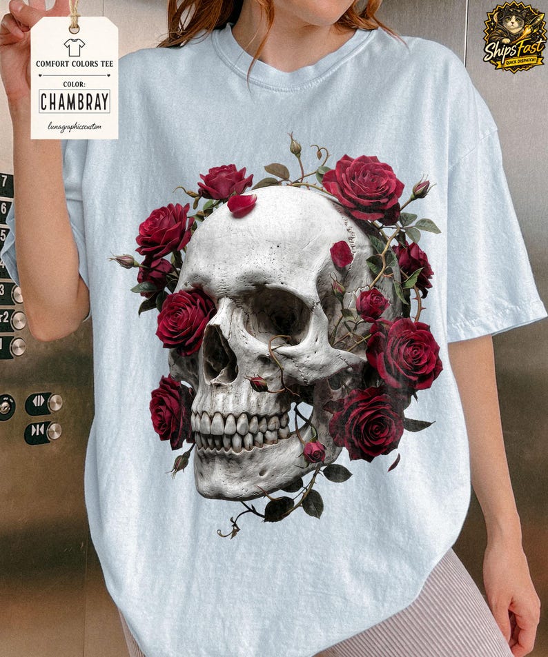 May include: Light blue Comfort Colors tee featuring a detailed skull design encircled by vibrant red roses. The graphic tee includes the text "CHAMBRAY" on a tag. The design is a blend of gothic and floral elements.