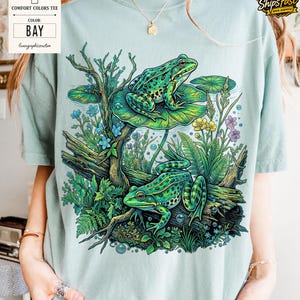 May include: A light green Comfort Colors tee shirt with a vibrant illustration of two green frogs on lily pads, surrounded by aquatic plants and flowers. The shirt's color is "Bay" as indicated on a tag.