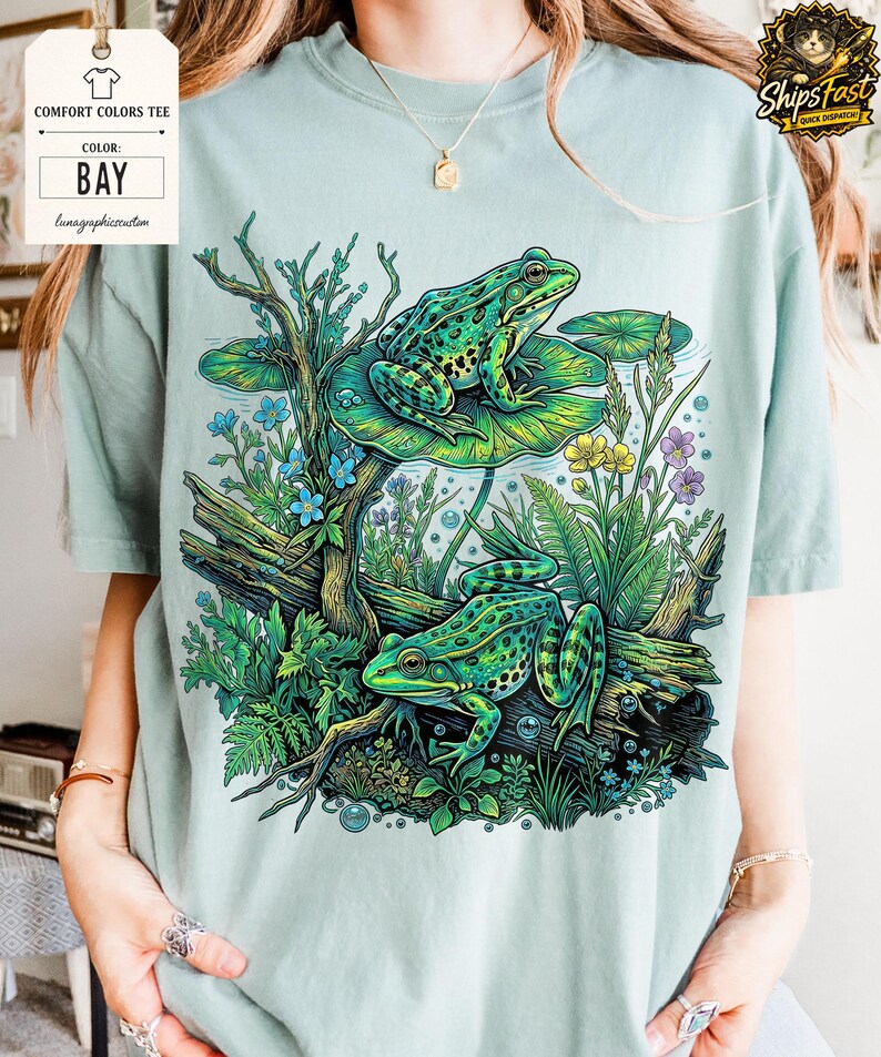 May include: A light green Comfort Colors tee shirt with a vibrant illustration of two green frogs on lily pads, surrounded by aquatic plants and flowers. The shirt's color is "Bay" as indicated on a tag.