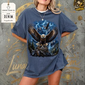 May include: Denim-colored Comfort Colors tee featuring a detailed graphic of a bald eagle with outstretched wings, set against a backdrop of mountains, lightning, and a waterfall. The eagle is perched on a nest with eaglets. The tee has the text "Comfort Colors Tee" and "Denim".