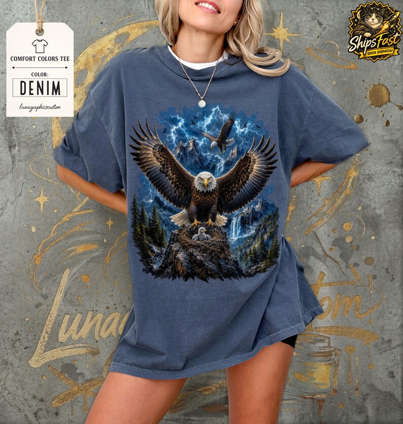 May include: Denim-colored Comfort Colors tee featuring a detailed graphic of a bald eagle with outstretched wings, set against a backdrop of mountains, lightning, and a waterfall. The eagle is perched on a nest with eaglets. The tee has the text "Comfort Colors Tee" and "Denim".