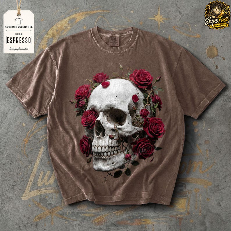 May include: Espresso-colored Comfort Colors tee featuring a detailed skull design encircled by red roses. The shirt has a vintage look, with the text "COMFORT COLORS TEE" and "ESPRESSO" on a tag.