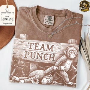 May include: A brown Comfort Colors T-shirt with a graphic of two monkeys and the words "TEAM PUNCH". The tee has a crew neck and short sleeves. A silver necklace with a pendant is draped across the shirt.