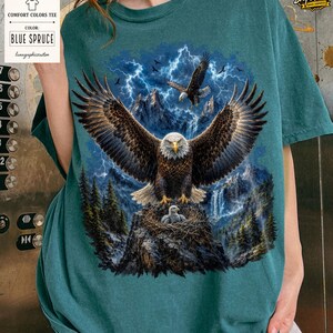 May include: Blue Spruce Comfort Colors tee featuring a detailed graphic of a bald eagle with outstretched wings, perched on a nest with eaglets. The design includes a mountain landscape, lightning, and the text "BLUE SPRUCE".