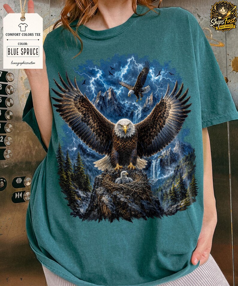 May include: Blue Spruce Comfort Colors tee featuring a detailed graphic of a bald eagle with outstretched wings, perched on a nest with eaglets. The design includes a mountain landscape, lightning, and the text "BLUE SPRUCE".