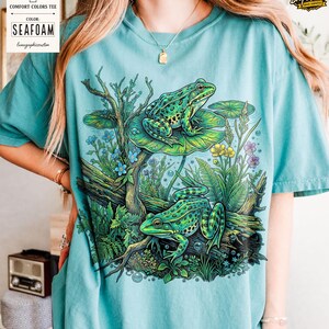 May include: Seafoam green Comfort Colors tee featuring a detailed illustration of two green frogs amidst aquatic plants and lily pads. The design includes blue, yellow, and purple flowers, and the text "Color: Seafoam" is visible on the upper left.