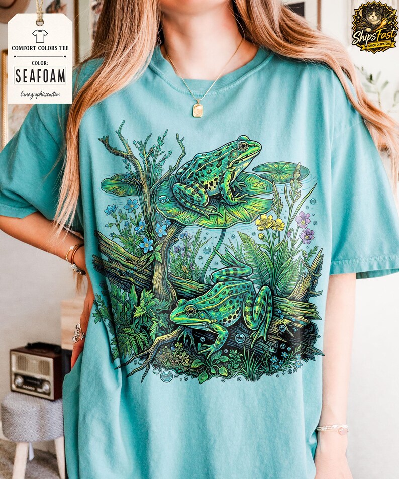 May include: Seafoam green Comfort Colors tee featuring a detailed illustration of two green frogs amidst aquatic plants and lily pads. The design includes blue, yellow, and purple flowers, and the text "Color: Seafoam" is visible on the upper left.