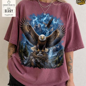 May include: Berry-colored Comfort Colors tee featuring a detailed graphic of an eagle with outstretched wings, perched on a rocky outcrop with a nest. The design includes a waterfall, mountains, and lightning.
