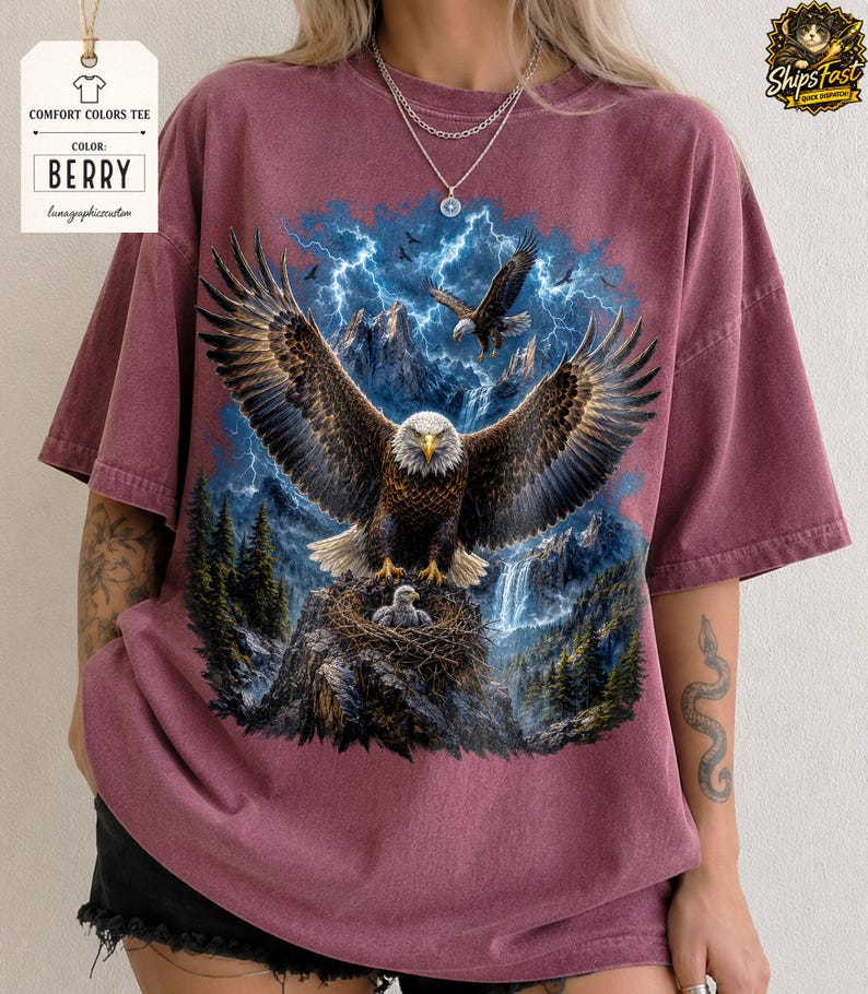 May include: Berry-colored Comfort Colors tee featuring a detailed graphic of an eagle with outstretched wings, perched on a rocky outcrop with a nest. The design includes a waterfall, mountains, and lightning.