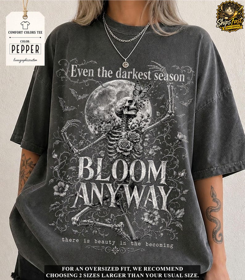 May include: A charcoal gray oversized t-shirt featuring a white graphic print of a skeleton surrounded by roses, a butterfly, and a full moon. The text reads: Even the darkest season, BLOOM ANYWAY, and there is beauty in the becoming.