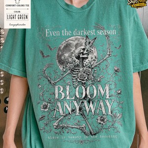 May include: A light green t-shirt featuring a graphic print of a skeleton surrounded by roses, a full moon, and bats. The text reads: Even the darkest season, BLOOM ANYWAY, there is beauty in the becoming. The design is rendered in black and white.