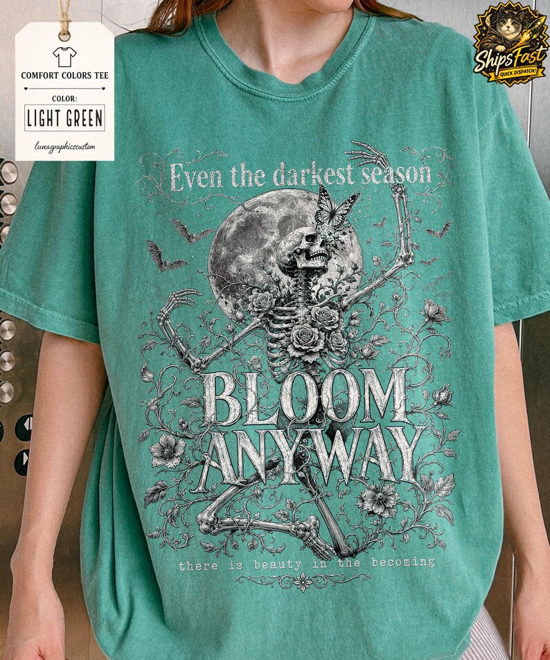 May include: A light green t-shirt featuring a graphic print of a skeleton surrounded by roses, a full moon, and bats. The text reads: Even the darkest season, BLOOM ANYWAY, there is beauty in the becoming. The design is rendered in black and white.