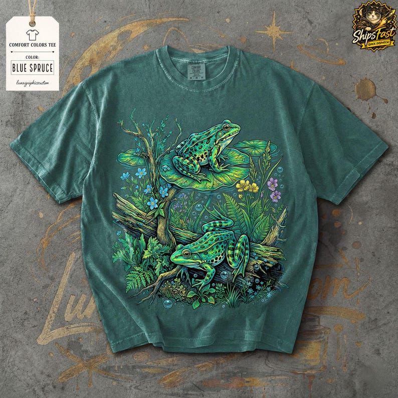 May include: Blue Spruce Comfort Colors tee featuring a vibrant illustration of two green frogs amidst lush aquatic plants and flowers. The design includes the text "Comfort Colors Tee" and "Blue Spruce" on a tag.