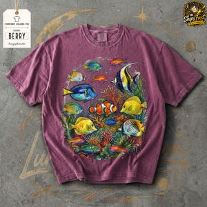 May include: Berry-colored Comfort Colors tee featuring a vibrant illustration of various colorful fish, coral, and aquatic plants. The design includes a clownfish, blue tang, and other tropical fish. The tag reads "Comfort Colors Tee" and "Berry".