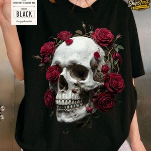 May include: Black Comfort Colors tee featuring a detailed skull design encircled by vibrant red roses. The skull has realistic shading and is the focal point of the graphic. The tag indicates the shirt color is black.