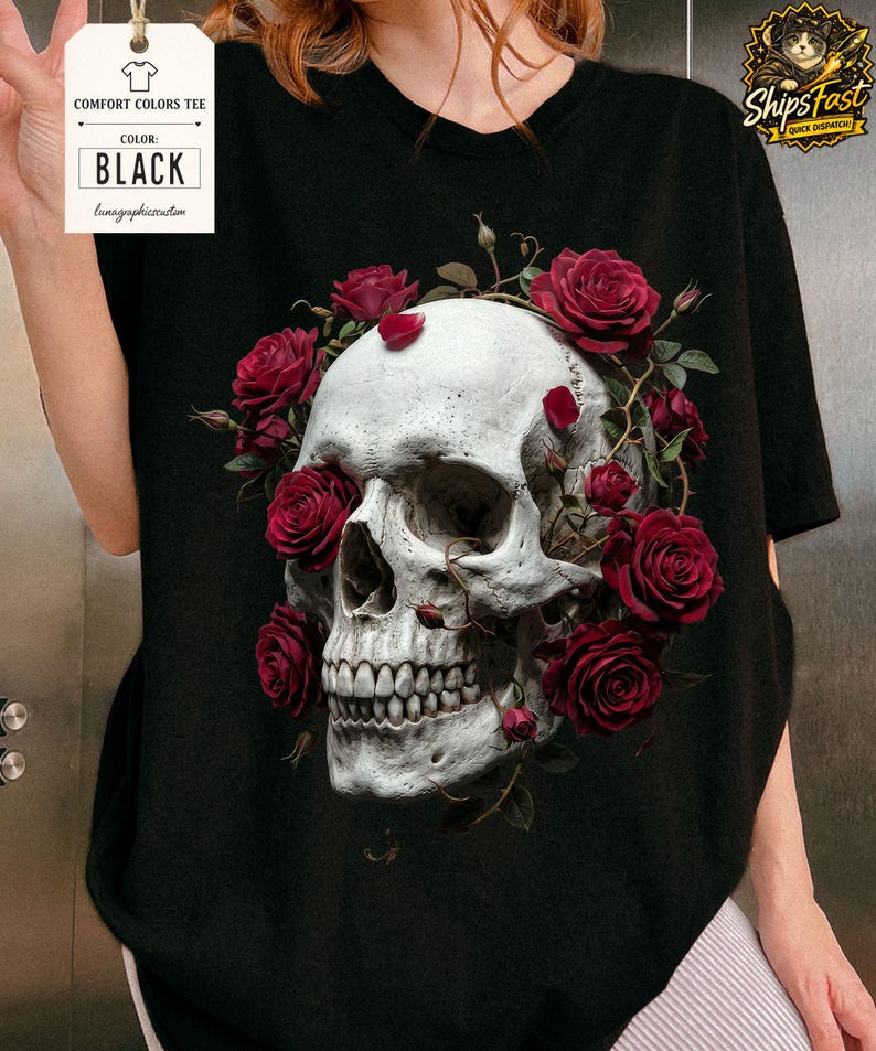 May include: Black Comfort Colors tee featuring a detailed skull design encircled by vibrant red roses. The skull has realistic shading and is the focal point of the graphic. The tag indicates the shirt color is black.